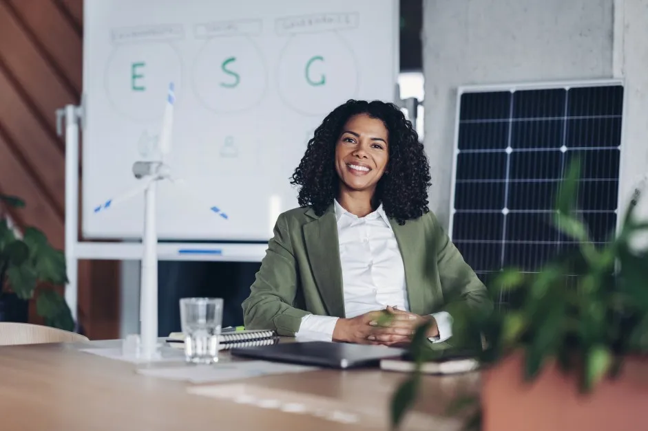 Mulher empresária sorri no escritório estando de frente para uma placa solar, representando os benefícios da energia solar para empresas