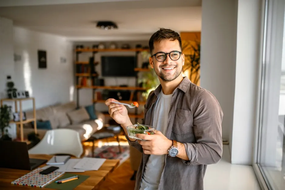 Homem come uma salada feliz enquanto olha pela janela se questionando se pode ter energia solar no seu apartamento